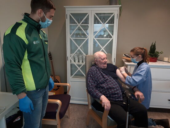 Bilde fra vaksinasjon på Liabø. Kristian Korsnes får vaksine av turnuslege Anne Grethe Gulla. Sykehjemslege Harald Grimstad bistår