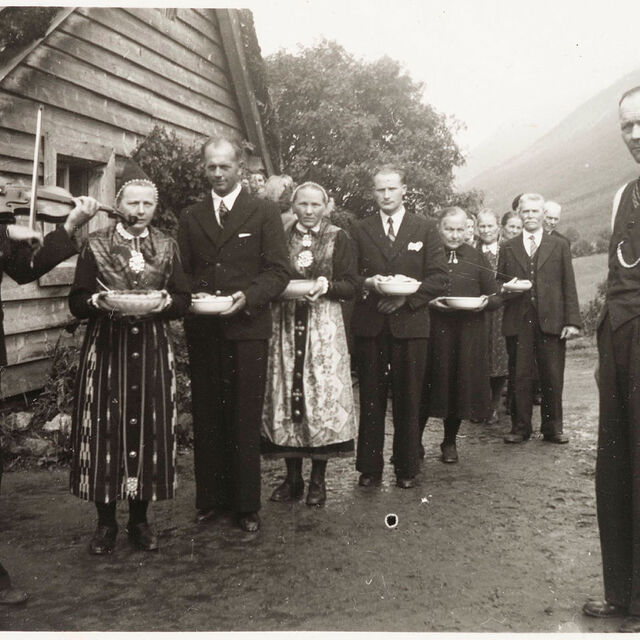 svart-kvitt-bilde av ei gruppe folk, menn i dress, damer i bunad, spelemann med fele til høgre.