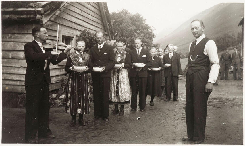 svart-kvitt-bilde av ei gruppe folk, menn i dress, damer i bunad, spelemann med fele til høgre.