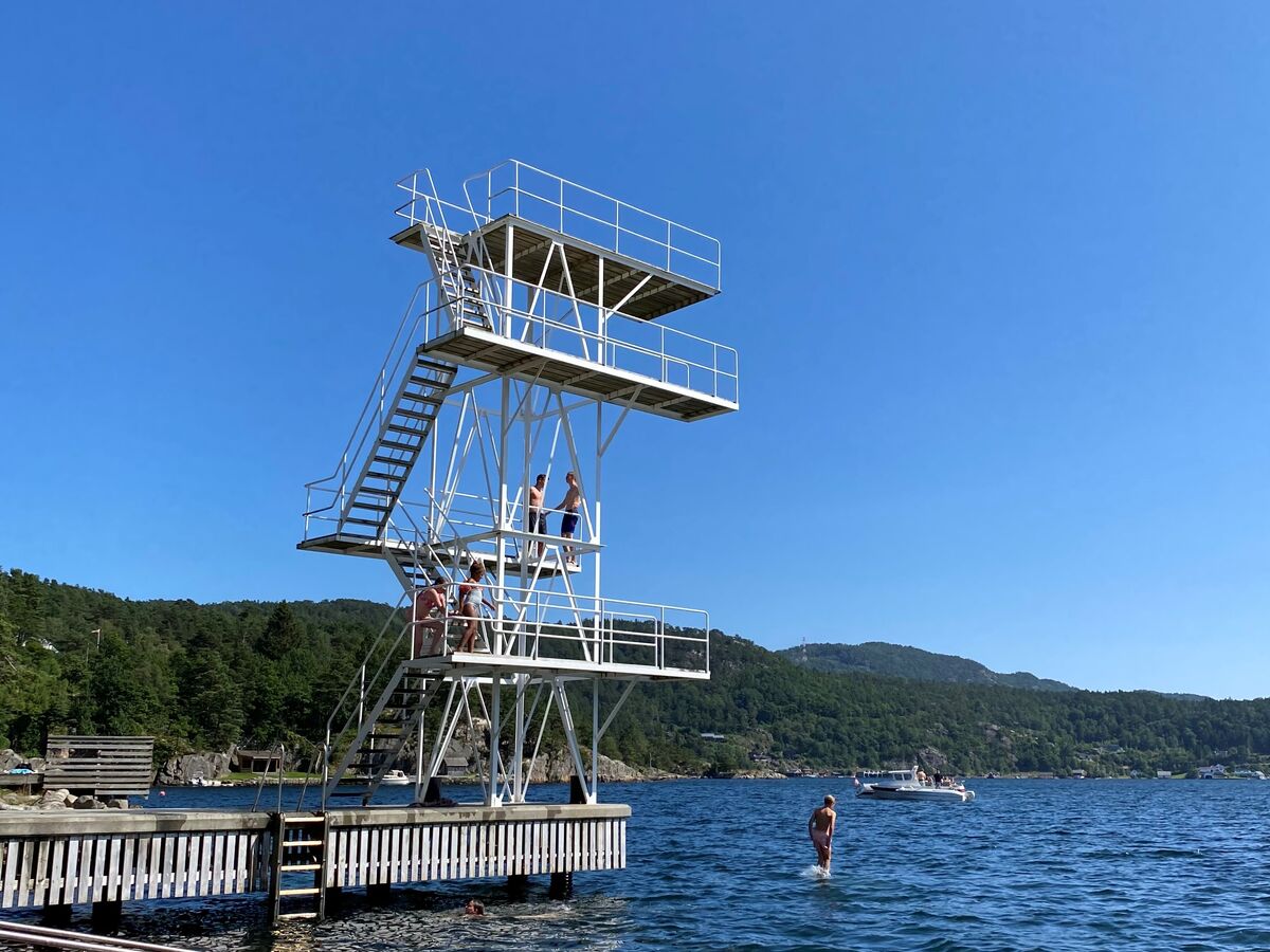 Stupetårnet på Grønnes i midten av bildet, omringet av hav og strand. Blå himmel. Enkelte mennesker i bakgrunnen.