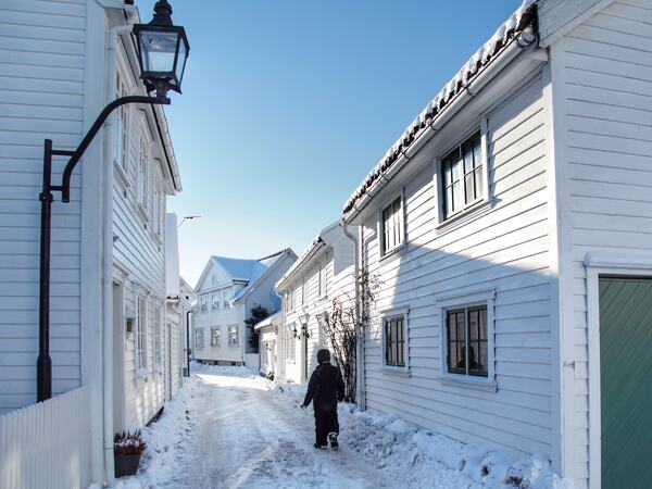 Person som går i snødekt gate full av hvite sørlandshus. Blå himmel.