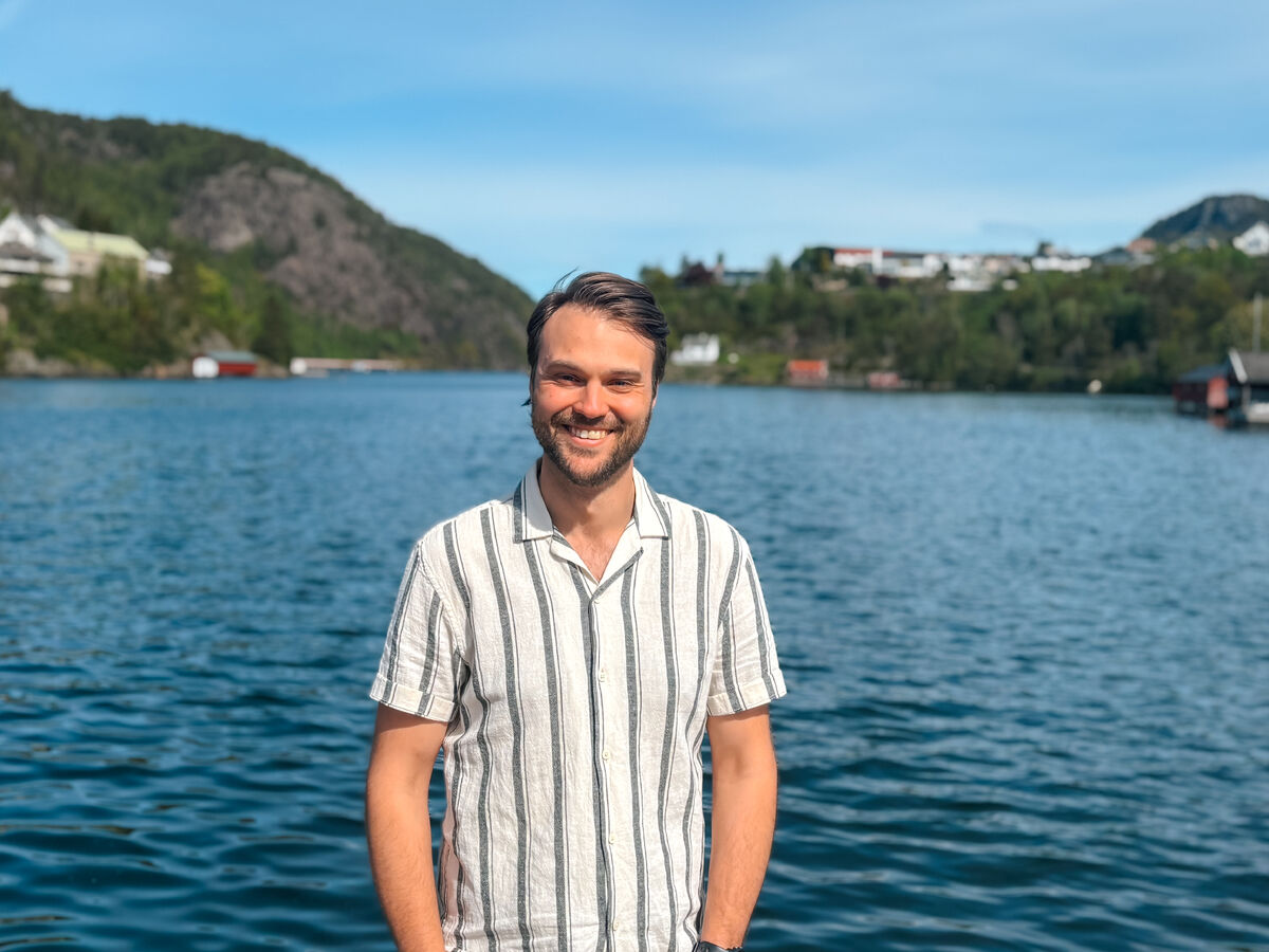 Hvit mann med mørkt hår og stripete skjorte, blå himmel og sjø som bakgrunn.
