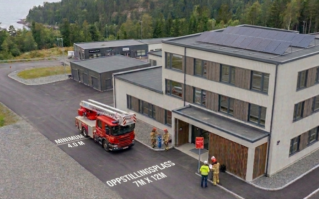 Luftfoto av en rød brannbil foran en treetasjes næringsbygning med solcelletak. På asfalten står det «MINIMUM 4.0 M» og «OPPSTILLINGSPLASS 7M X 12M». Fire personer i refleksvester står ved en blå hydrant og et lite, rødt skilt nær inngangen.